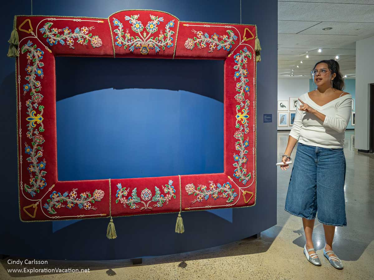 A woman standing by a giant frame with traditional Metis beadwork.